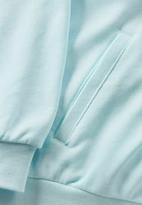 Tessuto per felpa azzurro chiaro con una trama liscia, caratterizzato da una tasca laterale e polsini a costine. Le cuciture sono uniformi e pulite.