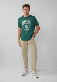 Cyan t-shirt met witte grafische ontwerpen, beige broek en grijs-witte sneakers met zwarte accenten. Casual pasvorm en een ronde halsontwerp.