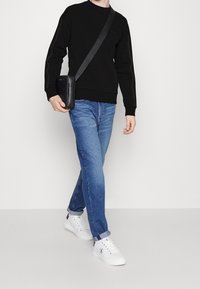 Felpa nera con polsini a coste, jeans blu con una leggera sfumatura e sneakers bianche. Una piccola borsa a tracolla nera è indossata sulla spalla.