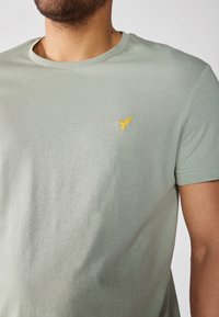 Camiseta de algodón verde claro con cuello redondo, que presenta un pequeño logotipo de un pájaro amarillo bordado en el lado izquierdo del pecho. Textura lisa.