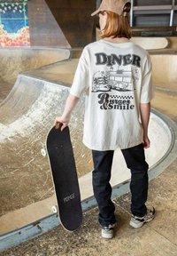 T-shirt blanche oversize avec un graphique de diner noir au dos, pantalon noir et planche à roulettes. Fond de skatepark en bois fermé.