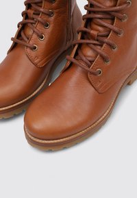 Las botas de tobillo de cuero marrón presentan una textura lisa, punta redonda, ojales de metal y cordones marrones. La suela de goma gruesa proporciona durabilidad.