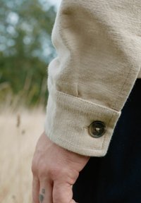 Beige corduroy overhemdmanchet met een donkere knoop. Hand die het manchet vasthoudt, waardoor de textuur en details van de stof zichtbaar zijn tegen een vage achtergrond.