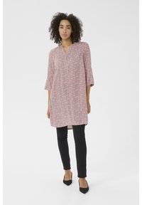 Tunique longue rose à motif en zigzag, décolleté en V et manches trois-quarts, associée à un pantalon noir slim et des chaussures noires à bout pointu.