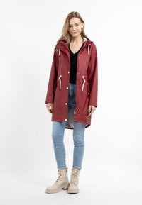 Giacca antipioggia rossa con cappuccio, dettagli bianchi, tasche frontali e chiusura con bottoni, abbinata a jeans skinny azzurri e stivali beige.