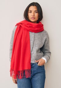 Écharpe en laine rouge élégamment drapée sur un cardigan gris en maille avec fermeture à boutons. Un jean en denim bleu complète la tenue. Détails en franges visibles sur l'écharpe.