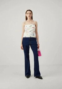 White tank top with multicolored playful patterns, paired with dark blue flared jeans. Model holds a small pink bag and wears black shoes.