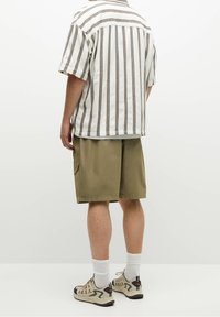 Camisa blanca de manga corta con rayas verticales marrones, combinada con pantalones cortos verde oliva. Modelo con zapatillas beige y calcetines blancos.