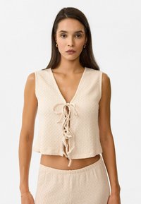 Beiger Cropped-Top mit strukturiertem Muster, V-Ausschnitt und vorderer Schnürung. Weicher Stoff mit lässiger Passform.