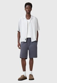 Camicia a maniche corte bianca a righe sottili, pantaloni grigi, canotta bianca e sabot marroni, caratterizzati da una vestibilità comoda e dettagli dei bottoni in evidenza.