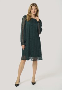 Teal lace dress with a diamond pattern, elbow-length sheer sleeves, a loose fit, and a hem that falls above the knee. Worn with black heels.