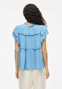 Blusa azzurro chiaro con un design a volant, maniche corte e un'apertura a chiave sul retro. Realizzata in un tessuto morbido con una vestibilità comoda e una struttura a strati.