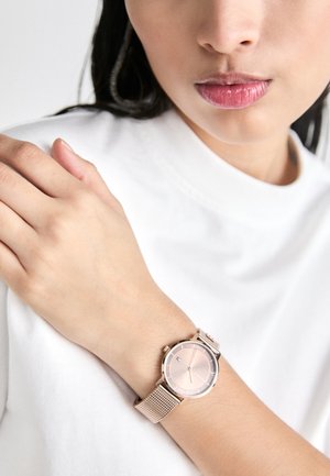 Round rose gold watch with mesh band, featuring a light pink dial and minimalist hour markers. Worn on a hand against a white fabric.