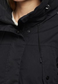 Svart väderbeständig jacka med hög krage, dragkedja och tryckknappar, justerbart snöre och slät, matt yta.