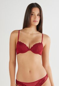 Roter Spitzen-BH mit gepolsterten Cups, gewellten Kanten, verstellbaren schmalen Trägern und einem passenden Spitzenbund an der Unterhose. Glatte Textur.