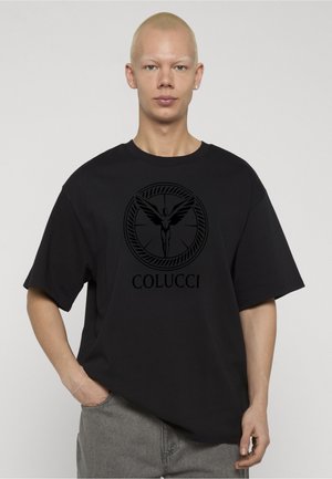 Person mit kurzen blonden Haaren, der ein schwarzes Colucci-T-Shirt mit Flügelfigur-Logo trägt und mit einer Hand in der Tasche einer grauen Hose steht.
