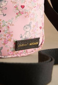Růžová taška s květinovými a Moominovými vzory, s tmavým uchem a značkovou nálepkou. Materiál se zdá být hladký s jemnou texturou.