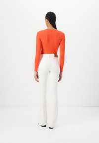 Suéter naranja de punto trenzado con mangas largas, combinado con jeans blancos acampanados de cintura alta que tienen bolsillos traseros y una etiqueta marrón en la cintura.