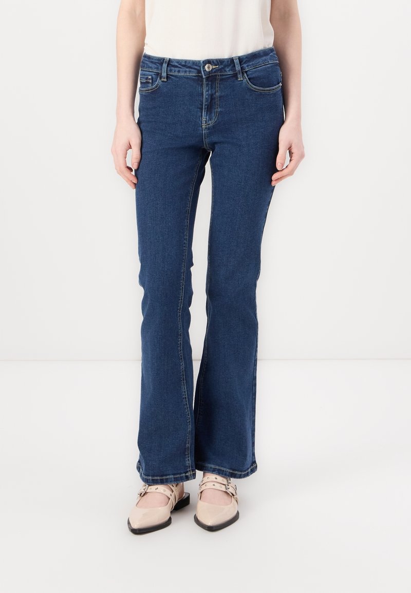 Femme portant un jean bootcut bleu foncé avec une taille boutonnée et des chaussures plates beiges à double bride.