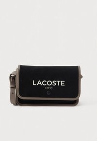 Black canvas crossbody bag with a tan trim, featuring a front flap, button closure, and white printed "LACOSTE 1933" logo.