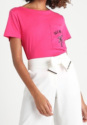 Femme portant un t-shirt rose vif avec un motif de palmier et un pantalon blanc taille haute doté d'une large ceinture nouée en nœud.