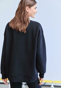 Čierna oversized mikina s okrúhlym výstrihom, rebrovanými manžetami a lemom, mäkkou textúrou a spustenými ramennými švami. Zobrazený pohľad zozadu.