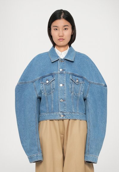 Donna che indossa una giacca di jeans blu oversize con maniche lunghe esagerate e pantaloni beige, su sfondo semplice.