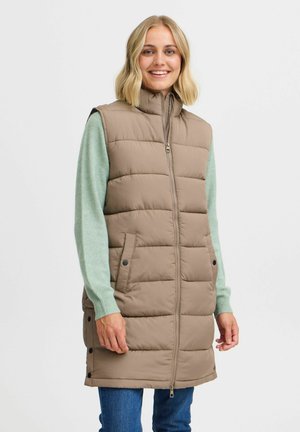 Gilet long rembourré de couleur brun clair avec un col haut, une fermeture éclair à l'avant et des poches latérales. Porté sur un pull vert, avec des détails à pression.