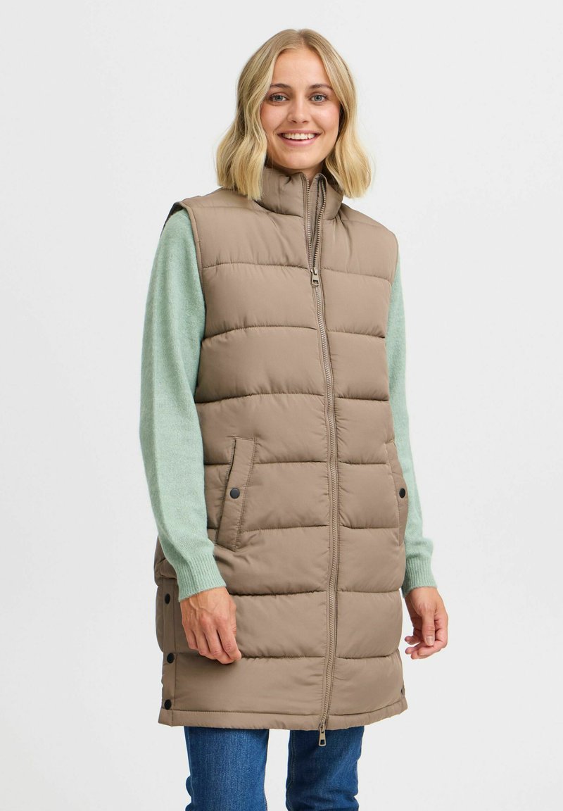 Gilet long rembourré de couleur brun clair avec un col haut, une fermeture éclair à l'avant et des poches latérales. Porté sur un pull vert, avec des détails à pression.