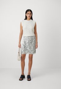 Haut sans manches en tricot blanc avec un motif texturé, associé à une jupe à franges argentées scintillantes. Chaussures à talons blocs ouvertes noires et petit sac.