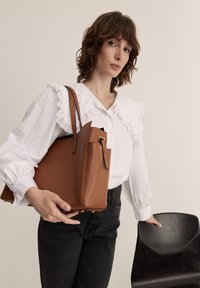 Sac cabas en cuir tan avec une forme structurée et des accents en métal doré. Le modèle le tient à côté d'une chaise en bois, portant un blouse blanche à volants.