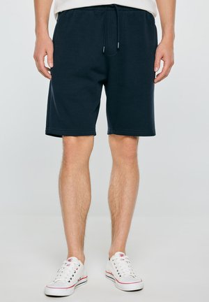 Mężczyzna w granatowych szortach na sznurki i białych niskich sneakersach, stoi na jednolitym jasnym tle.