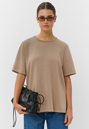 Femme portant un t-shirt beige oversize et des lunettes de soleil, tenant un sac en cuir noir avec cordon dans une main.