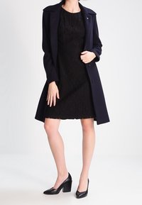 Schwarzes Spitzenkleid mit taillierter Silhouette, kombiniert mit einem dunkelblauen Mantel mit Kragen und schwarzen High Heels.