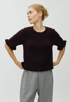 Femme aux cheveux blonds attachés en chignon, portant un pull sombre à manches courtes à volants et un pantalon gris, debout, les mains sur les hanches, regardant sur le côté.