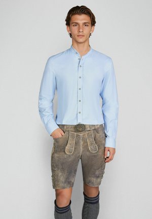 Jonge man die een lichtblauw overhemd draagt en traditionele bruine geborduurde leren korte broek met kniehoge sokken, staande tegen een effen achtergrond.