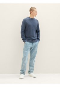 Szary sweter dzianinowy, luźne jasnoniebieskie dżinsy i białe sneakersy. Model stoi na tle jasnym, z rękami w kieszeniach.