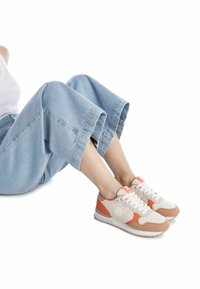 Zapatillas con una parte superior de malla beige, acentos de ante en color marrón claro y suela blanca. Presentan cordones y un logo circular en el lateral. Combinadas con pantalones de mezclilla claros y cortos.