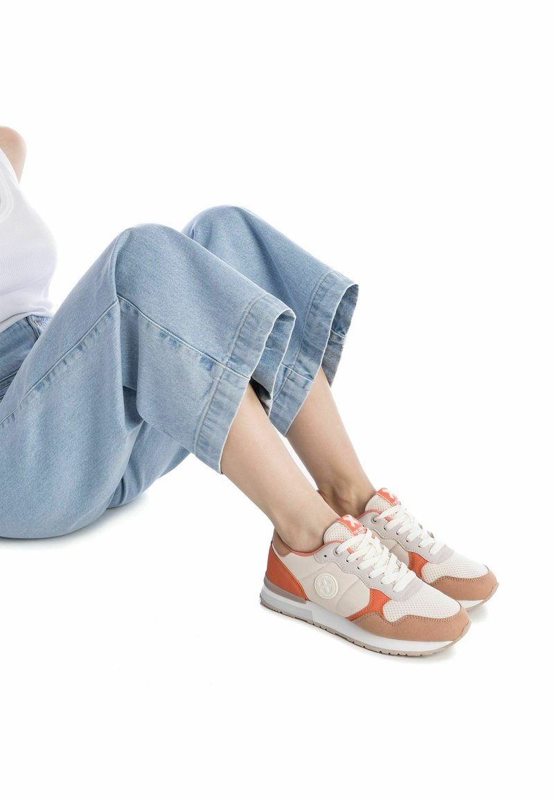 Zapatillas con una parte superior de malla beige, acentos de ante en color marrón claro y suela blanca. Presentan cordones y un logo circular en el lateral. Combinadas con pantalones de mezclilla claros y cortos.