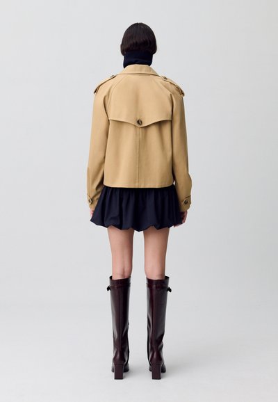Manteau beige avec un col rabattable, détails de boutonnage, porté sur une jupe noire. Des bottes brillantes marron jusqu'aux genoux complètent la tenue.