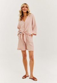 Světle růžový lněný dvoudílný outfit se zapínacím tričkem svázaným v pase a odpovídajícími šortkami s ohrnutými manžetami. Hnědé sandály.