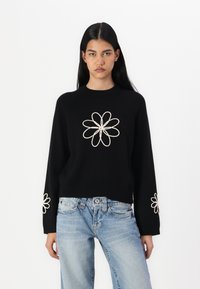 Svart strikket genser med lange ermer, med hvite broderte blomstermønstre på brystet og mansjettene. Kombinert med lyseblå jeans.