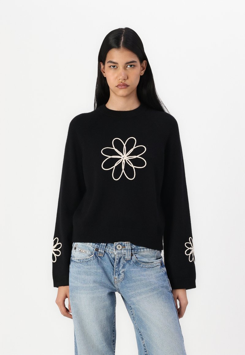Svart strikket genser med lange ermer, med hvite broderte blomstermønstre på brystet og mansjettene. Kombinert med lyseblå jeans.
