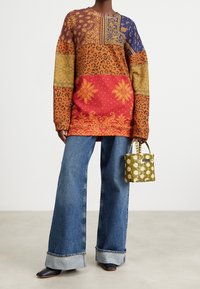 Camiseta de manga larga colorida con patrones mezclados en naranja, rojo, azul y marrón; combinada con jeans de pierna ancha azul claro y un bolso verde.