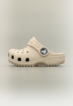 Zoccolo casual beige con fori di ventilazione, suola testurizzata e cinturino regolabile con un bottone a logo rotondo su sfondo neutro.