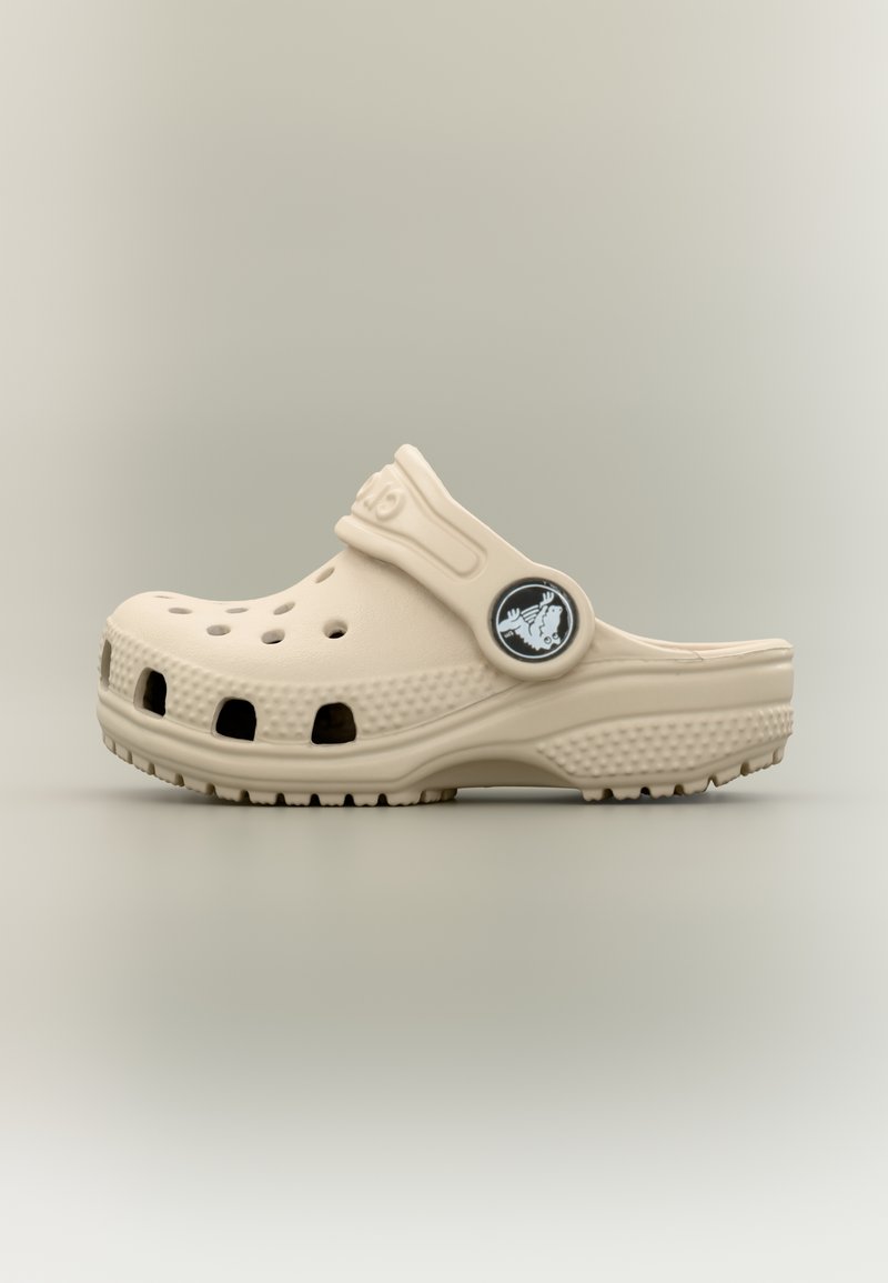 Μπεζ καθημερινό clog με οπές αερισμού, υφασμένο πέλμα και ρυθμιζόμενο λουράκι με στρογγυλό κουμπί λογότυπου σε ουδέτερο φόντο.