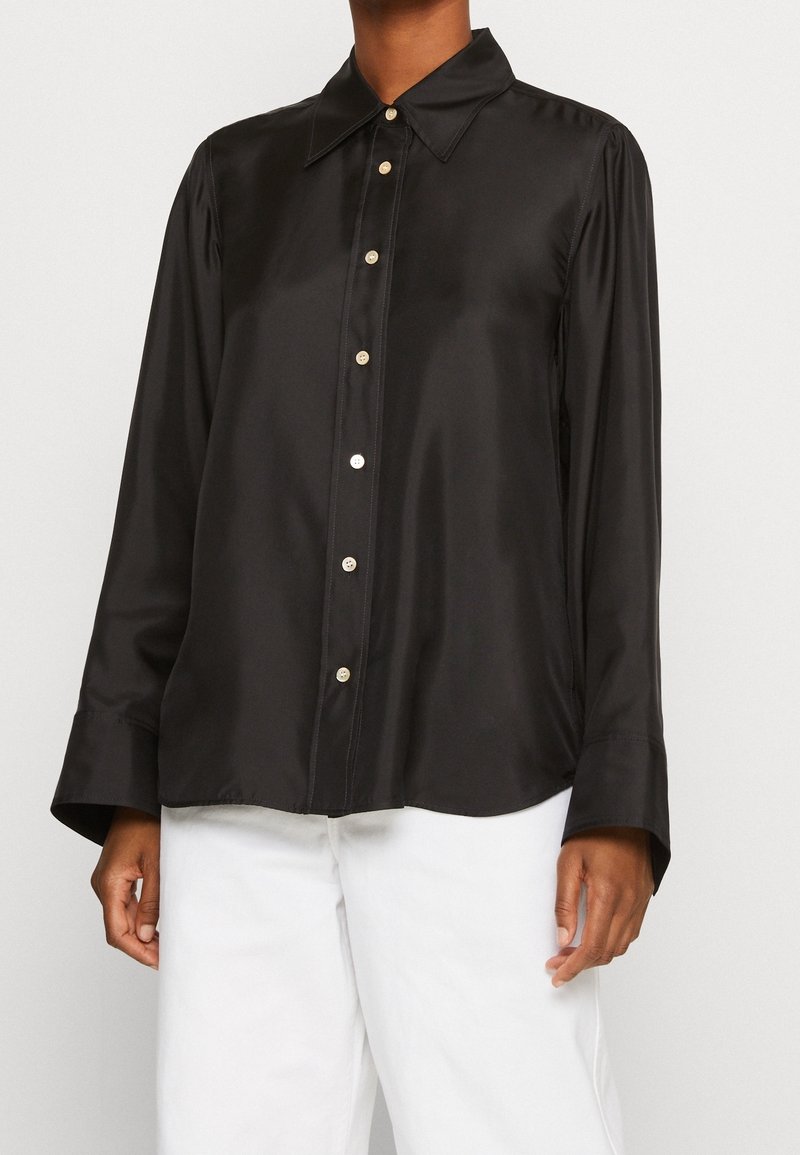 Personne portant une chemise noire en satin à manches longues boutonnée avec des boutons blancs, associée à un pantalon blanc, debout devant un fond uni.