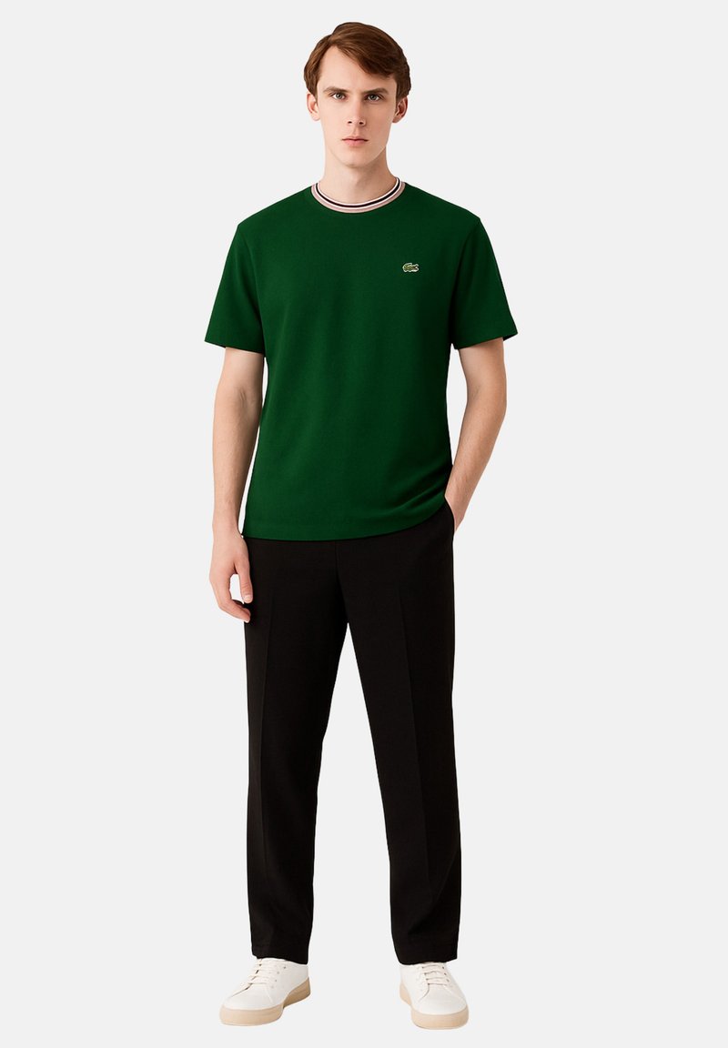 Camiseta verde de manga corta con escote redondo y ribete a rayas, combinada con pantalones negros y zapatillas blancas. Logo de Lacoste visible.