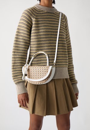Persoon draagt een gestreepte gebreide trui en een bruine geplooide rok, met een beige geweven handtas met witte bies en schouderriem.
