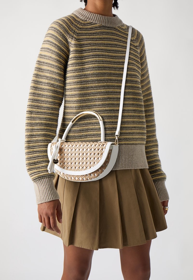 Personne portant un pull en tricot rayé et une jupe plissée marron, tenant un sac à main tissé beige avec bordure blanche et bandoulière.
